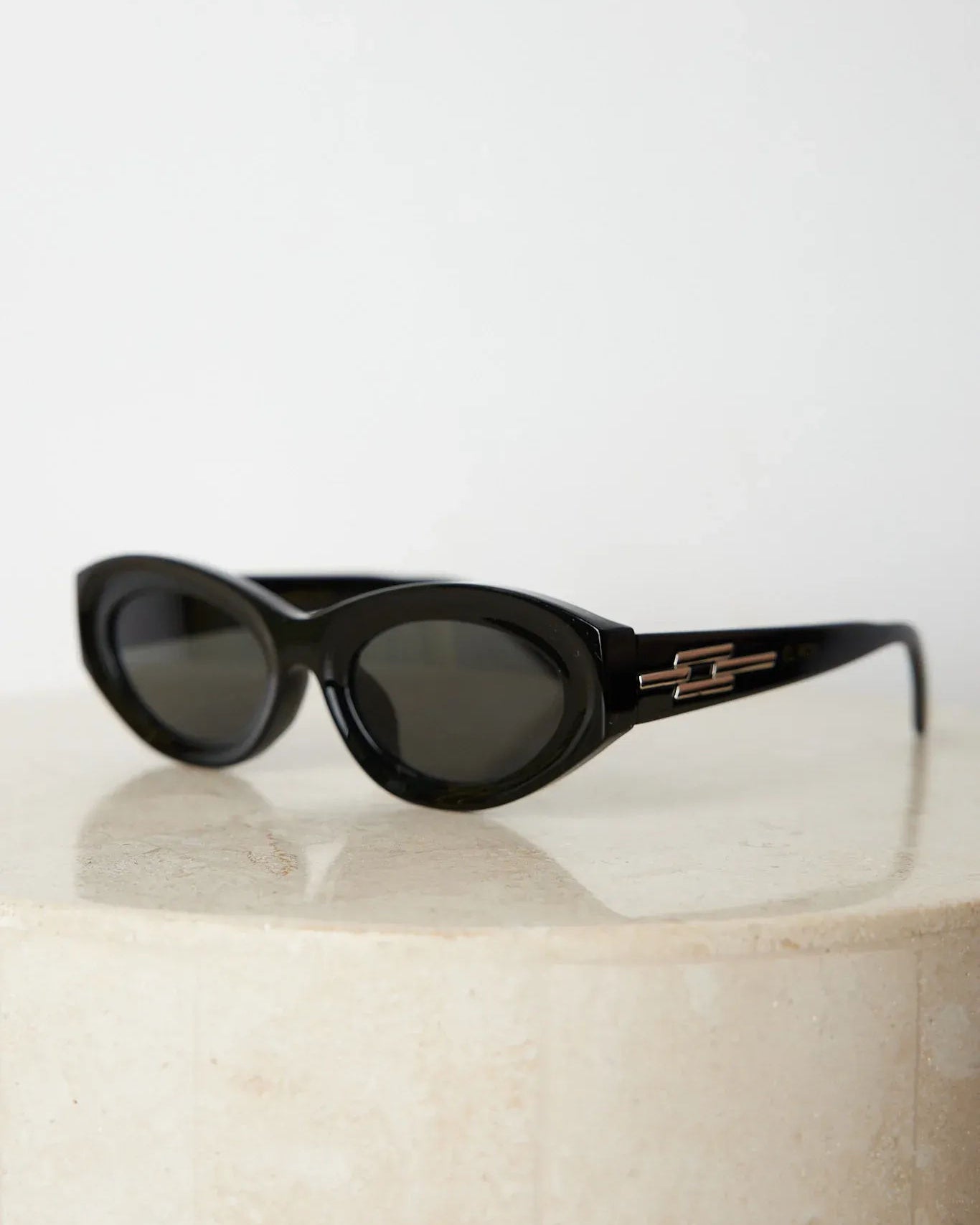 Close-up of a pair of black cat-eye sunglasses with dark lenses, resting on a smooth beige surface against a plain white background.