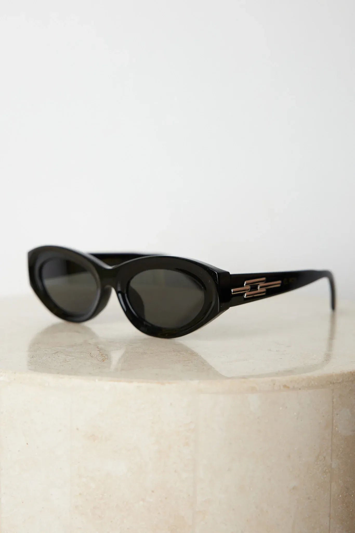 Close-up of a pair of black cat-eye sunglasses with dark lenses, resting on a smooth beige surface against a plain white background.