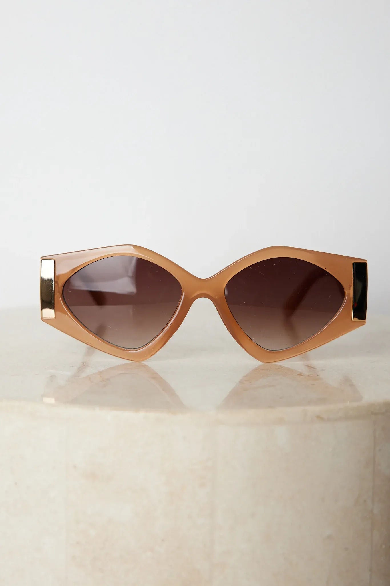 Brown cat-eye sunglasses with thick, glossy frames and dark brown tinted lenses, featuring gold accents on the corners, displayed on a light beige surface against a plain white background.