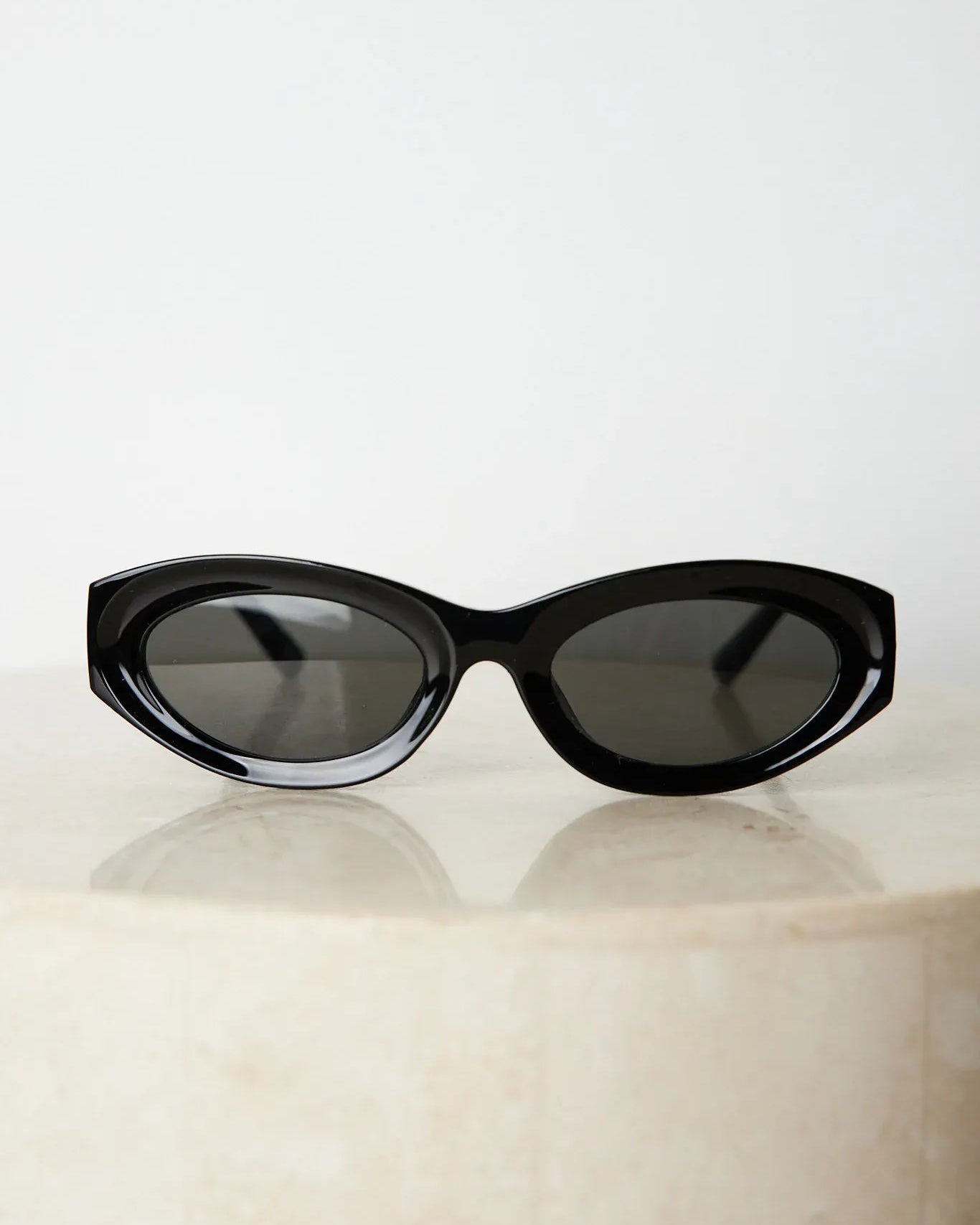 Close-up of a pair of black cat-eye sunglasses with dark lenses, resting on a smooth beige surface against a plain white background.