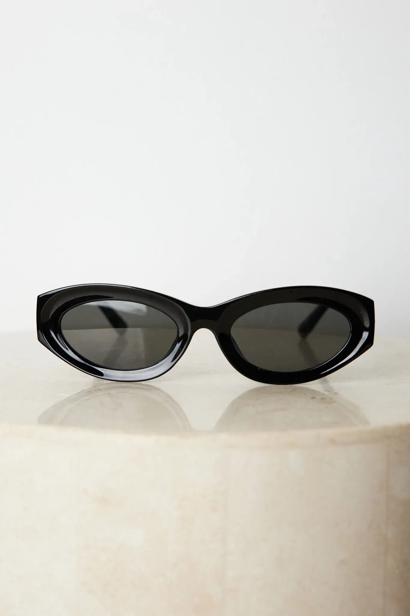 Close-up of a pair of black cat-eye sunglasses with dark lenses, resting on a smooth beige surface against a plain white background.