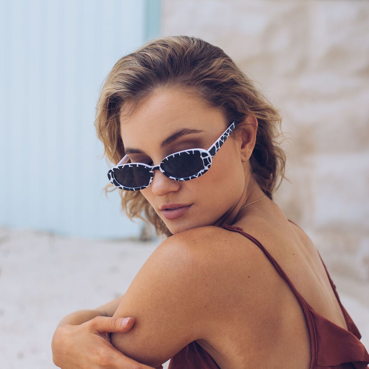Woman wearing narrow oval-shaped sunglasses with a bold black and white animal print frame, looking over her shoulder with a confident expression. She has wavy blonde hair and is dressed in a rust-colored spaghetti strap top, sitting against a soft beachy backdrop with sand, a light blue door, and a stone wall.