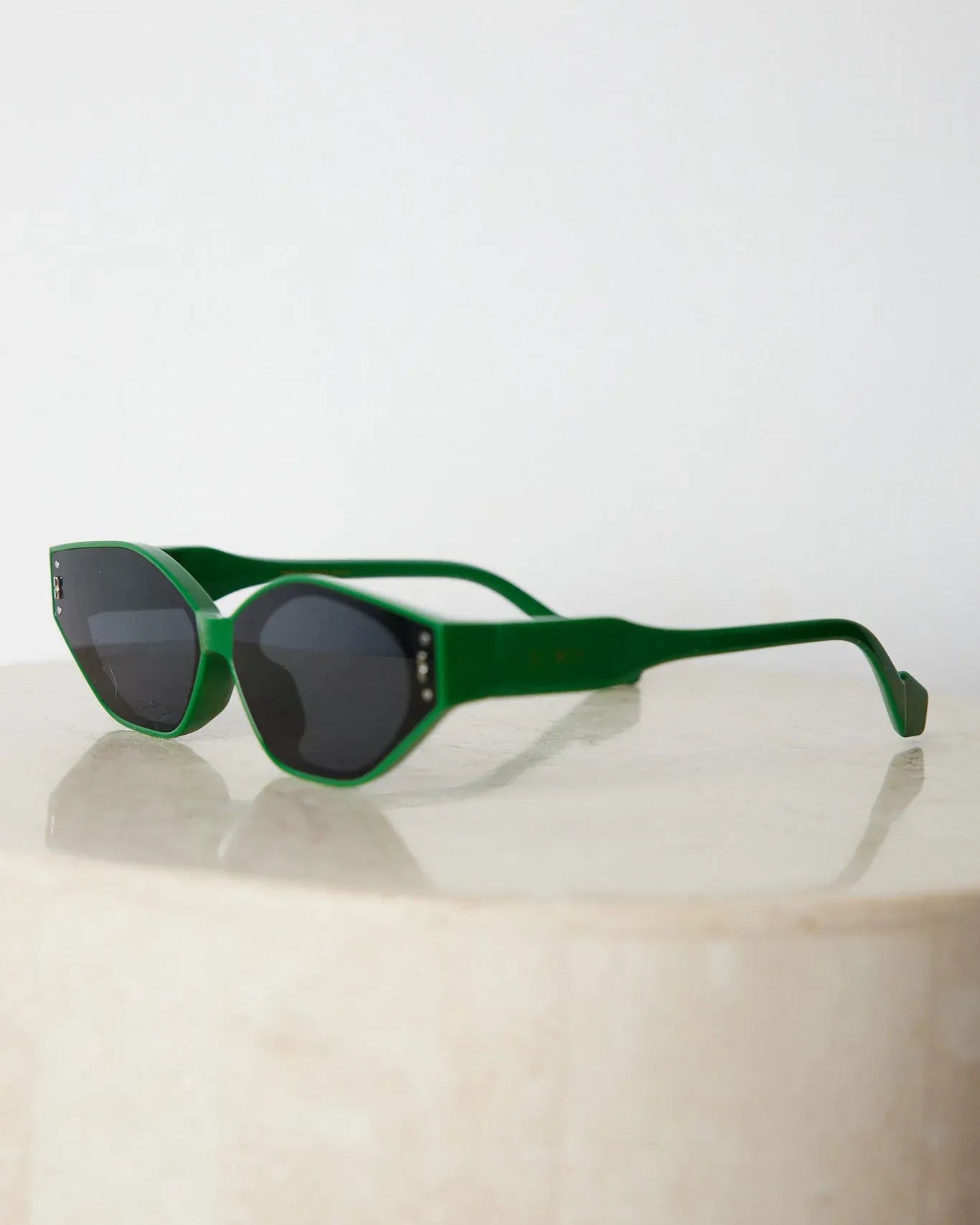 Green geometric-shaped sunglasses with dark lenses resting on a light marble surface against a plain white background.