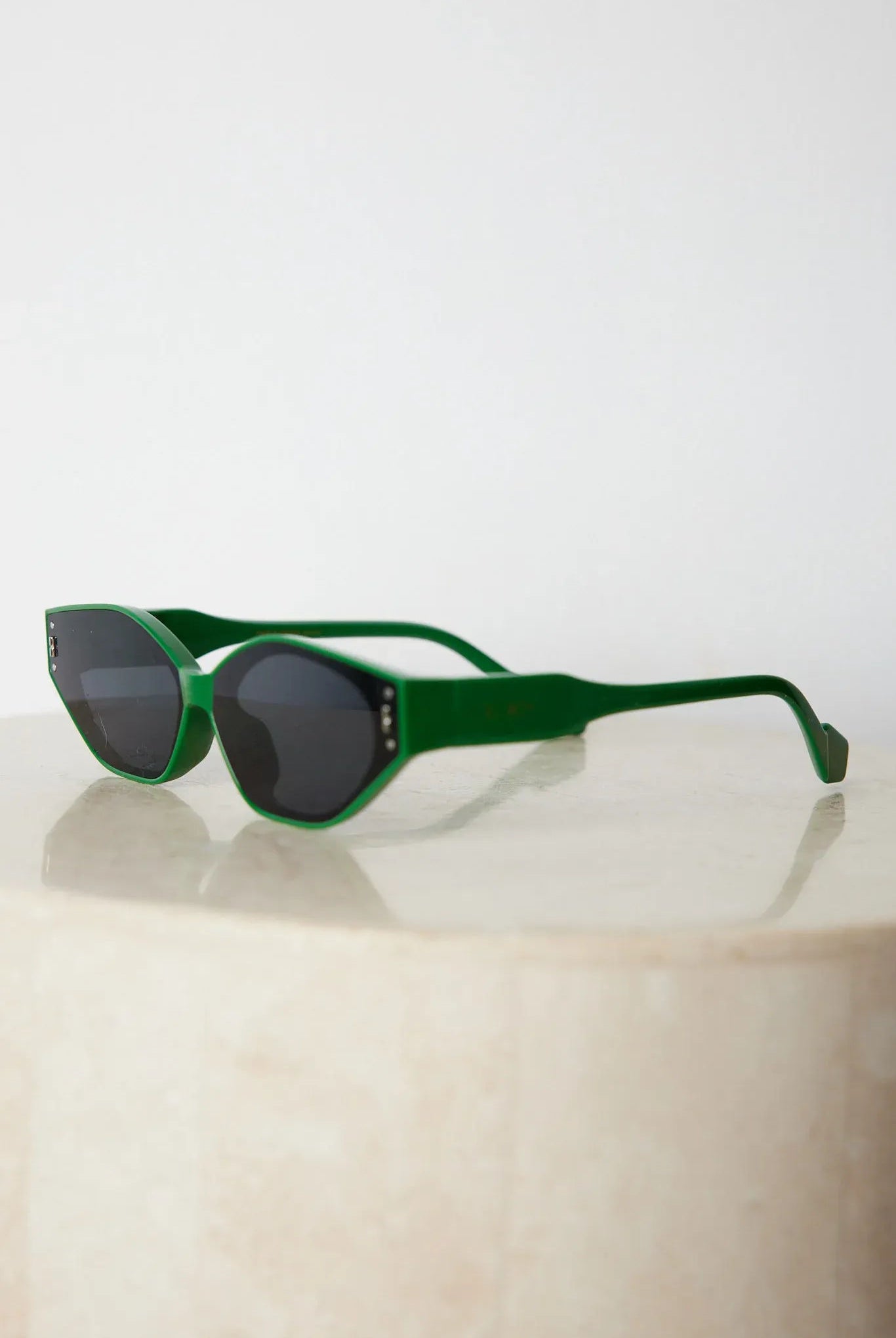 Green geometric-shaped sunglasses with dark lenses resting on a light marble surface against a plain white background.