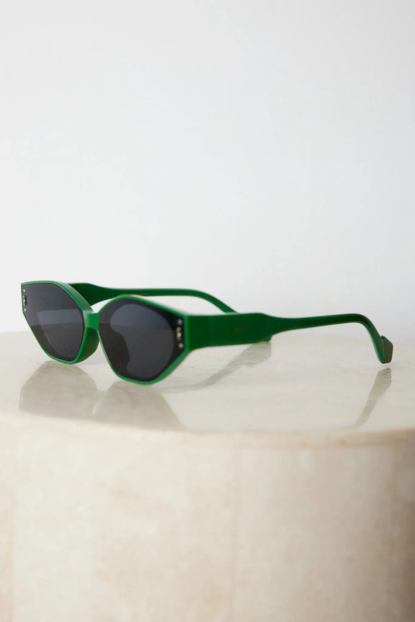 Green geometric-shaped sunglasses with dark lenses resting on a light marble surface against a plain white background.