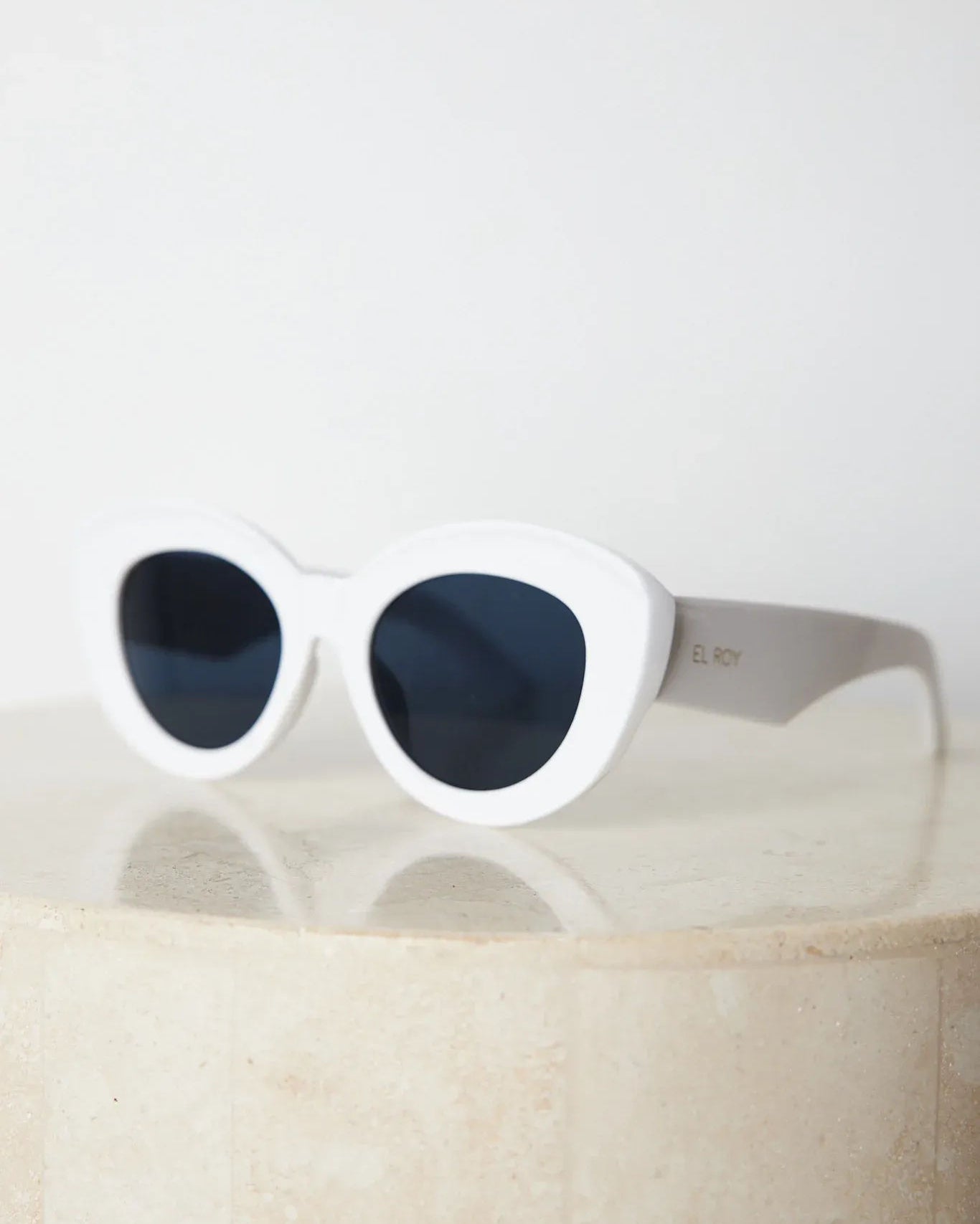 White cat-eye sunglasses with dark lenses displayed on a light beige marble surface against a plain white background.