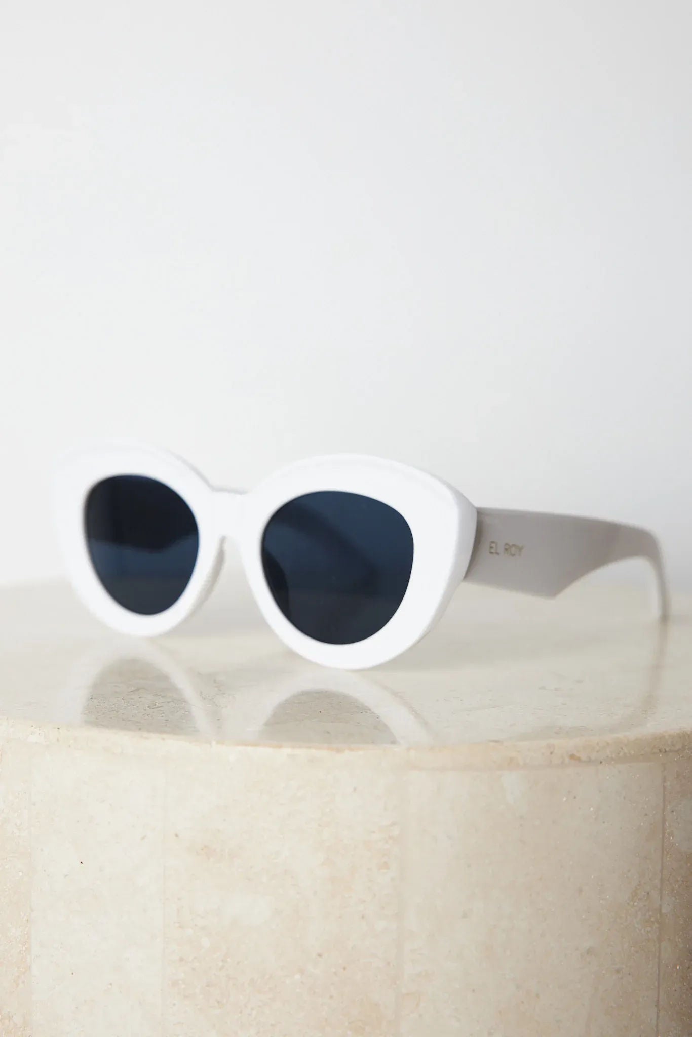 White cat-eye sunglasses with dark lenses displayed on a light beige marble surface against a plain white background.