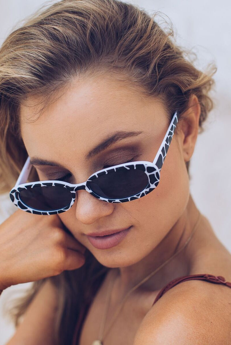 Woman wearing narrow oval-shaped sunglasses with a bold black and white animal print frame, looking over her shoulder with a confident expression. She has wavy blonde hair and is dressed in a rust-colored spaghetti strap top, sitting against a soft beachy backdrop with sand, a light blue door, and a stone wall.