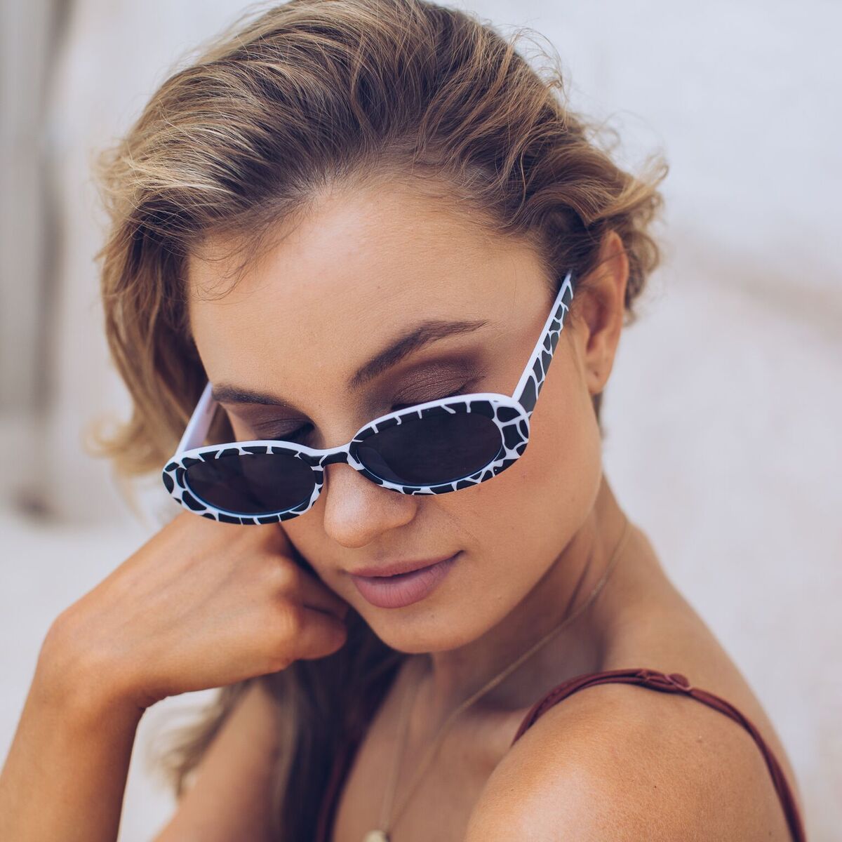 Woman wearing narrow oval-shaped sunglasses with a bold black and white animal print frame, looking over her shoulder with a confident expression. She has wavy blonde hair and is dressed in a rust-colored spaghetti strap top, sitting against a soft beachy backdrop with sand, a light blue door, and a stone wall.