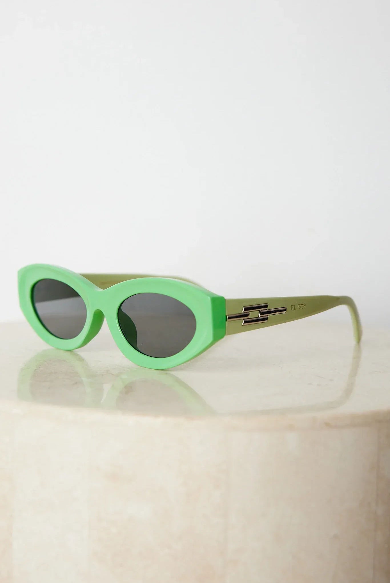 Close-up of a pair of bright green cat-eye sunglasses with dark lenses, placed on a smooth beige surface with a plain white background.