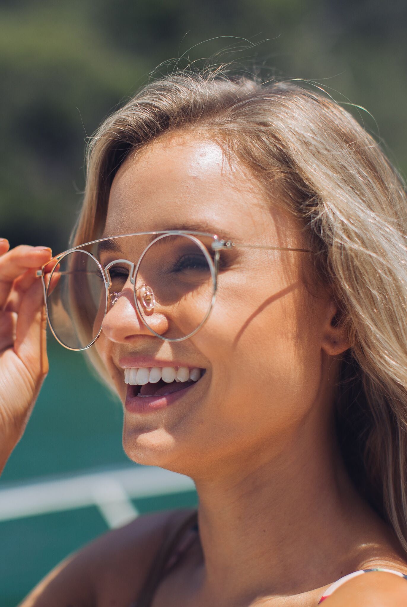Close-up of a hand holding a pair of gold-framed round eyeglasses with a double bridge and black temple tips. The background shows a light wooden floor and a woman’s legs in white shorts and beige open-toe heels, slightly out of focus.