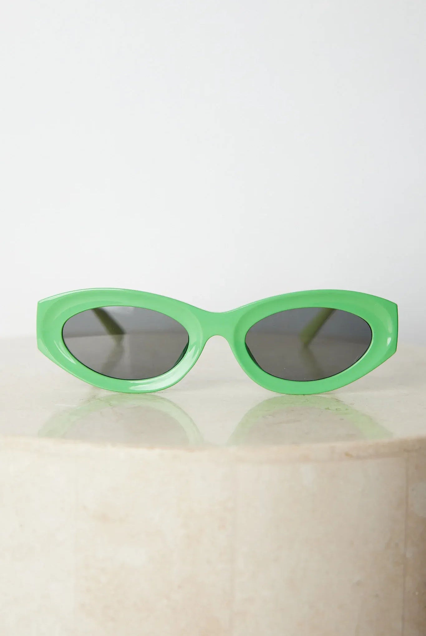 Close-up of a pair of bright green cat-eye sunglasses with dark lenses, placed on a smooth beige surface with a plain white background.