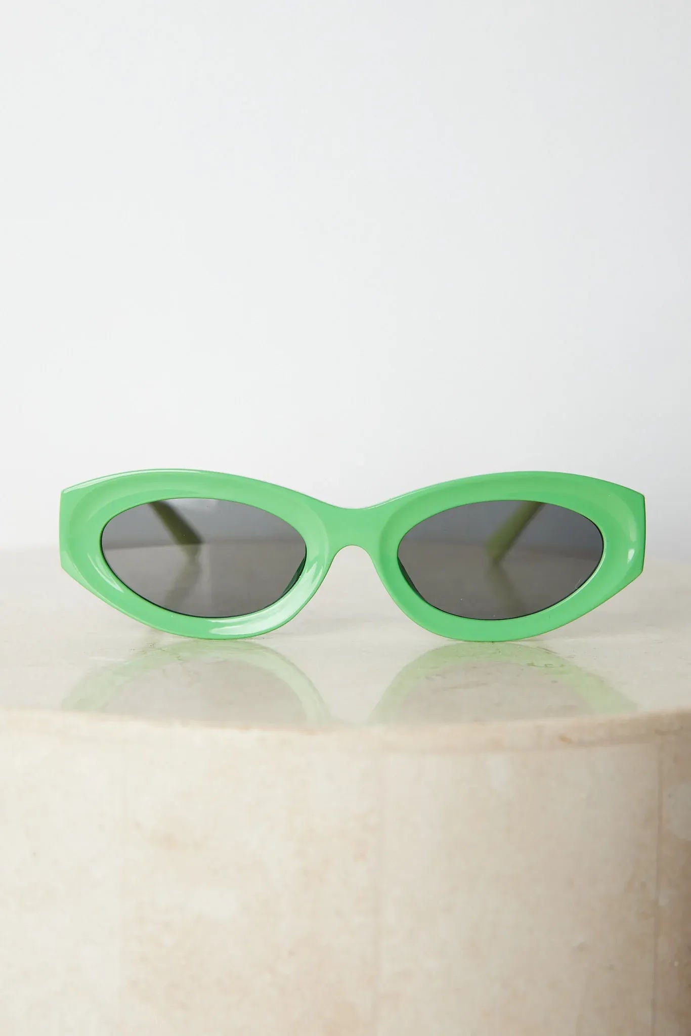 Close-up of a pair of bright green cat-eye sunglasses with dark lenses, placed on a smooth beige surface with a plain white background.