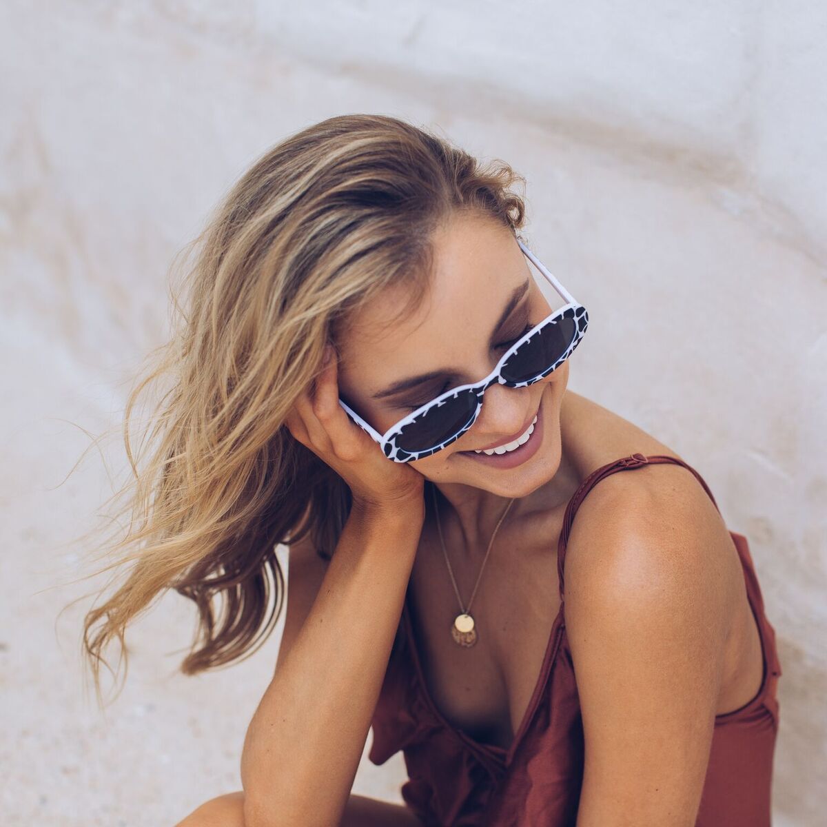 Woman wearing narrow oval-shaped sunglasses with a bold black and white animal print frame, looking over her shoulder with a confident expression. She has wavy blonde hair and is dressed in a rust-colored spaghetti strap top, sitting against a soft beachy backdrop with sand, a light blue door, and a stone wall.