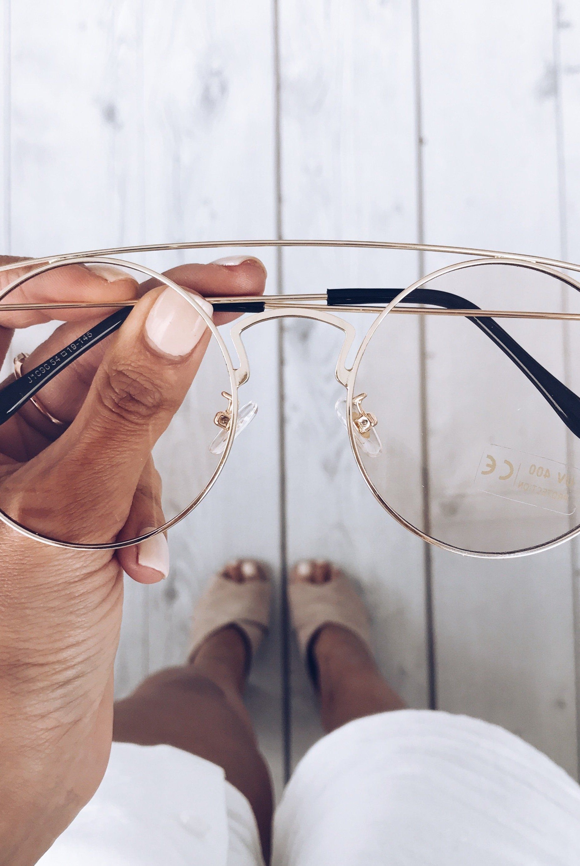 Close-up of a hand holding a pair of gold-framed round eyeglasses with a double bridge and black temple tips. The background shows a light wooden floor and a woman’s legs in white shorts and beige open-toe heels, slightly out of focus.