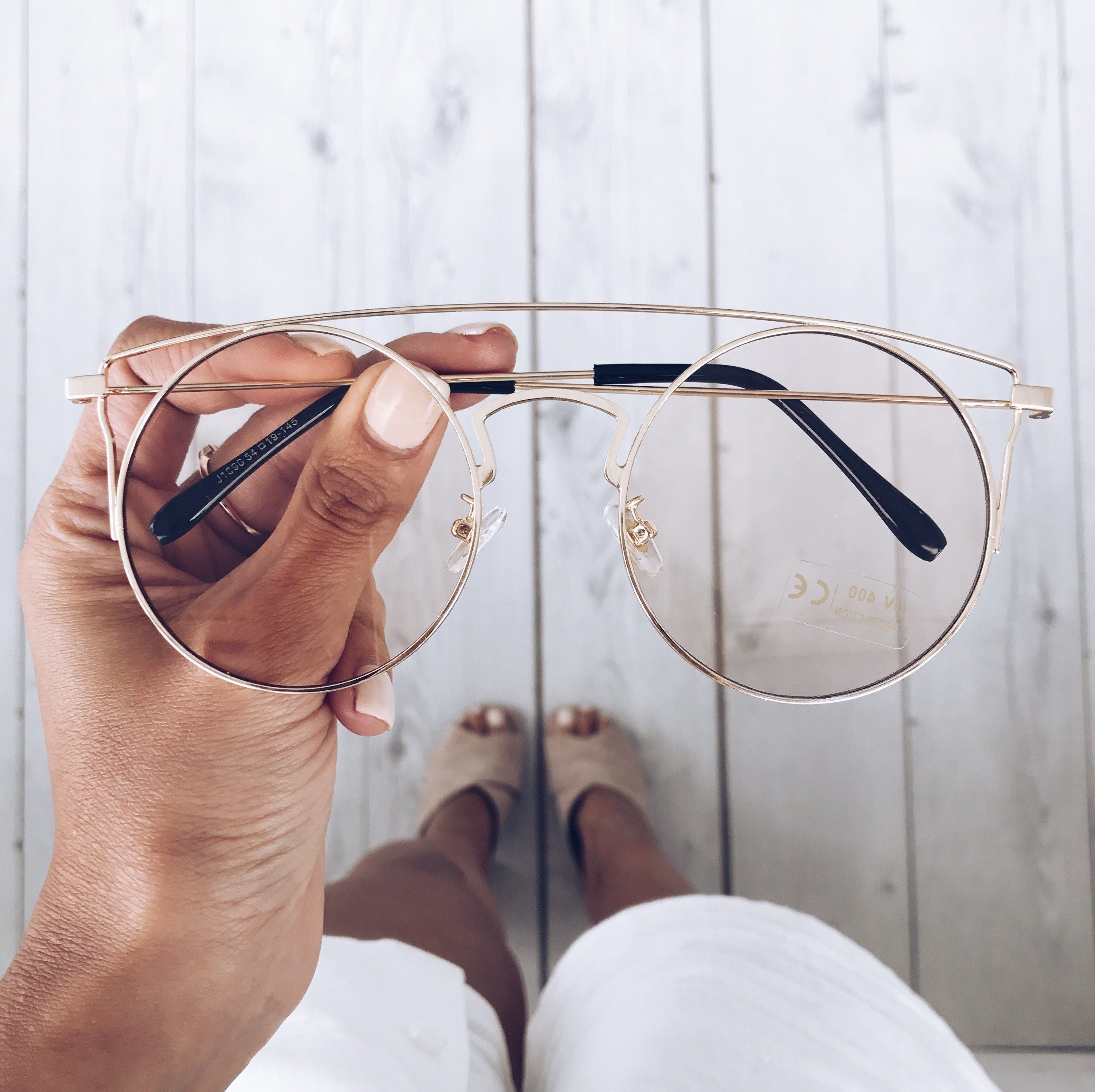 Close-up of a hand holding a pair of gold-framed round eyeglasses with a double bridge and black temple tips. The background shows a light wooden floor and a woman’s legs in white shorts and beige open-toe heels, slightly out of focus.