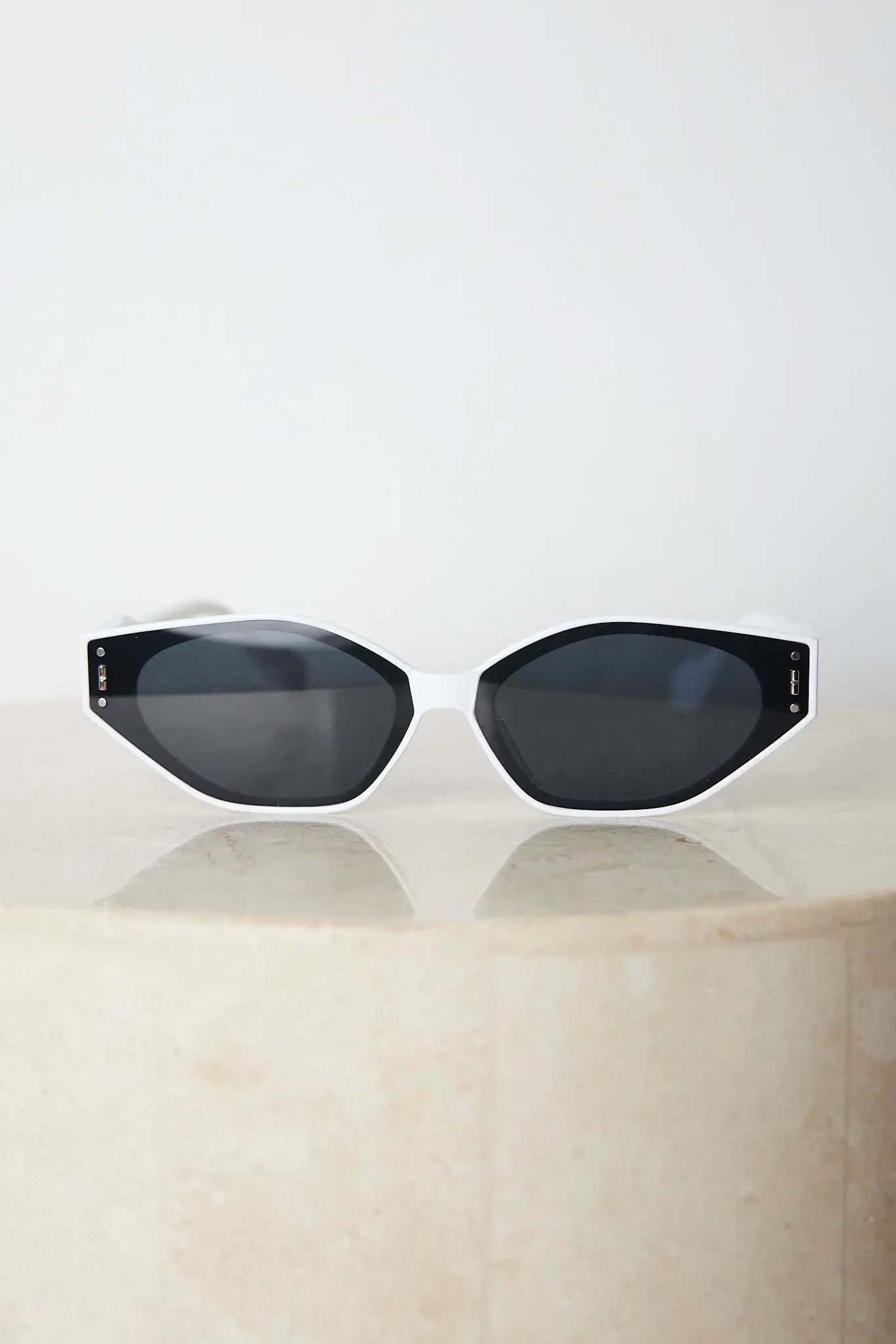 White geometric-shaped sunglasses with dark lenses resting on a light marble surface against a plain white background.