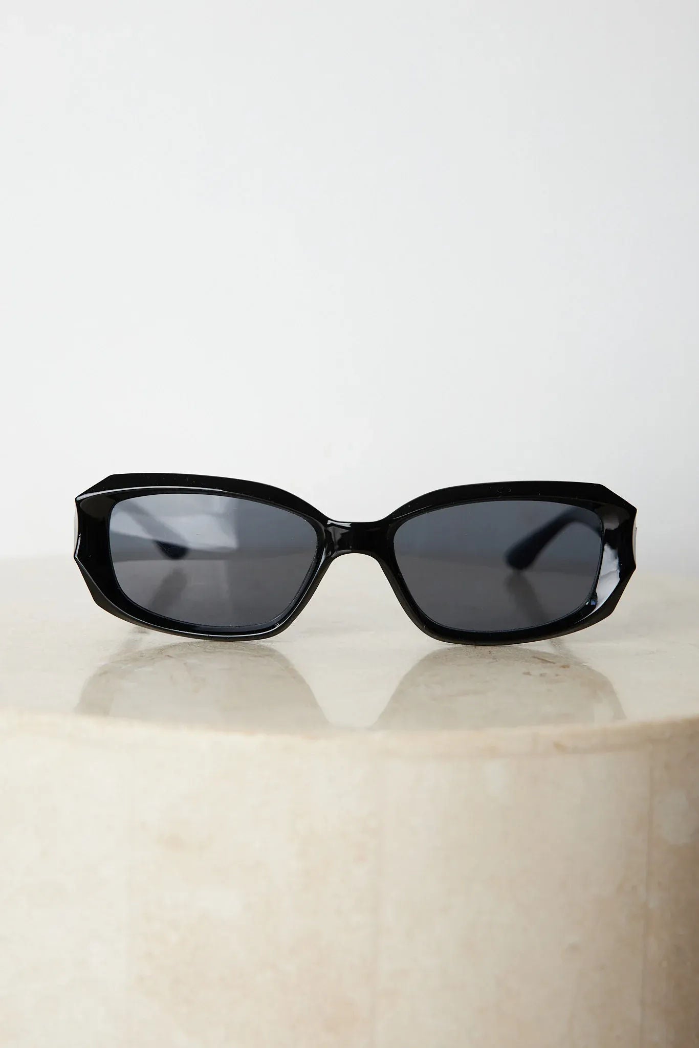 Black rectangular sunglasses with a glossy frame and dark tinted lenses, displayed on a light beige curved surface against a plain white background.