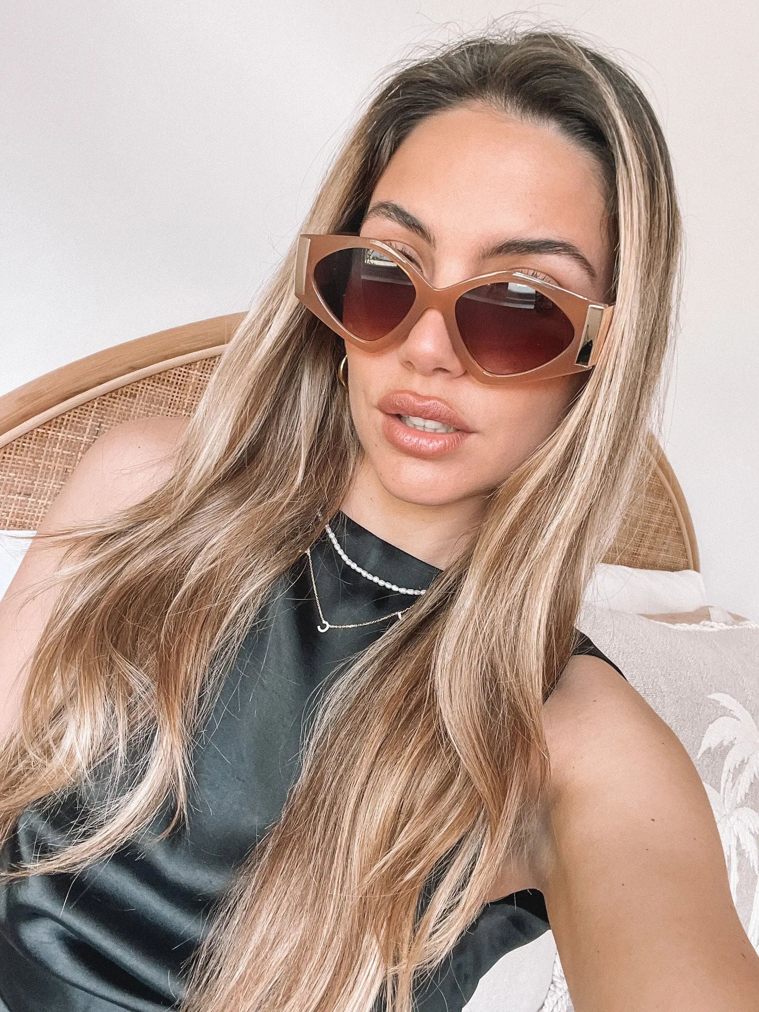 Brown cat-eye sunglasses with thick, glossy frames and dark brown tinted lenses, featuring gold accents on the corners, displayed on a light beige surface against a plain white background.