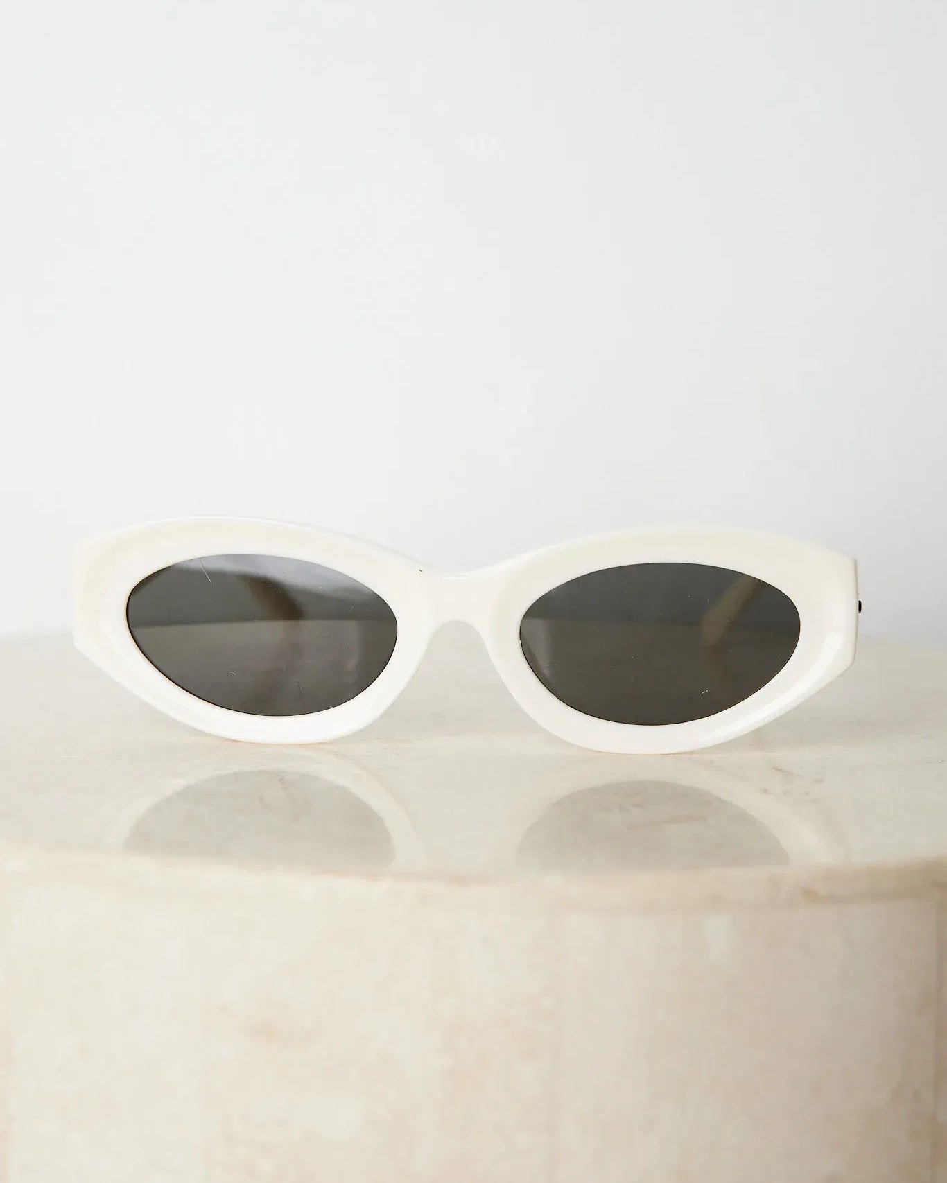 Close-up of a pair of white oval-shaped sunglasses with dark lenses, resting on a smooth beige surface against a plain white background.