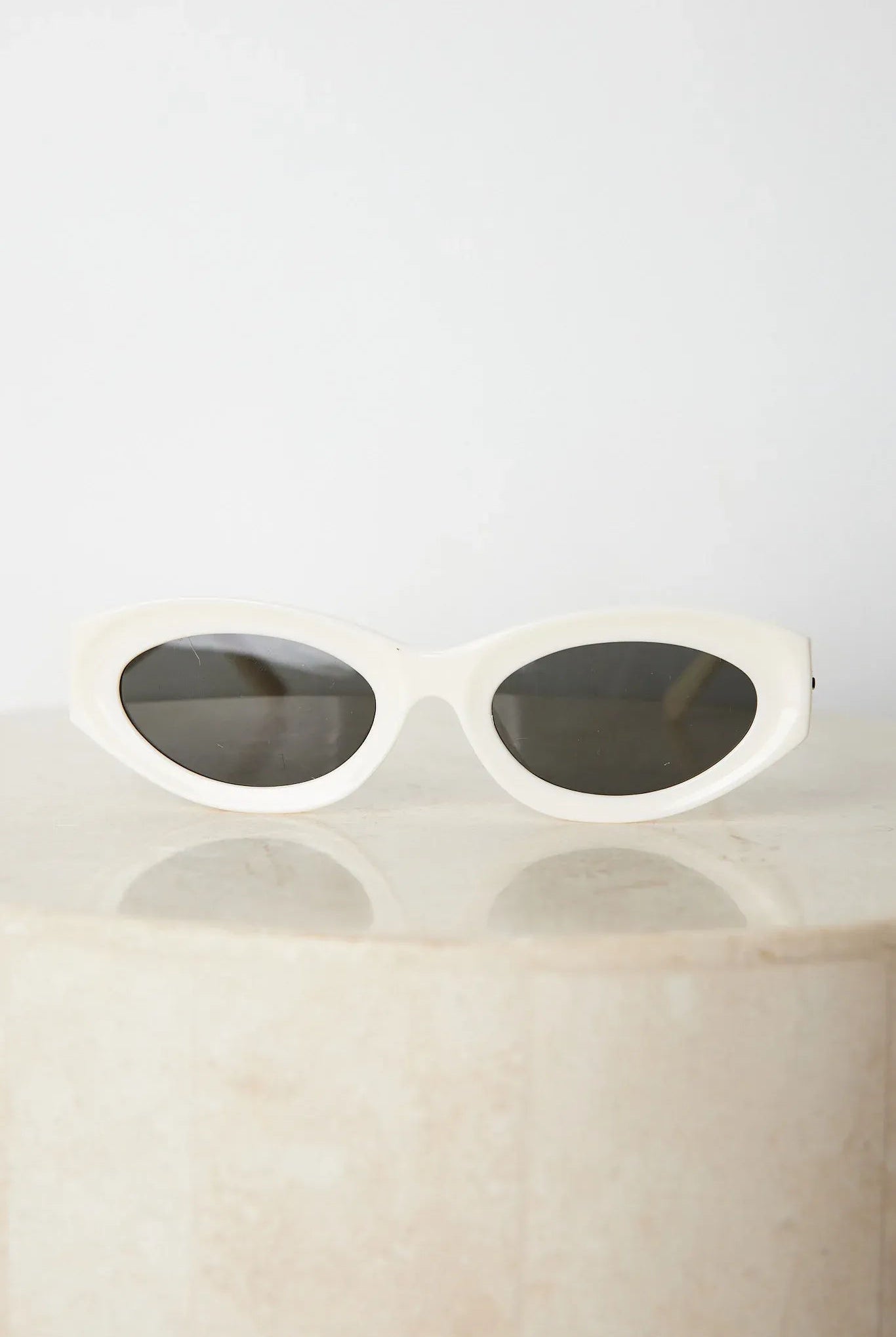Close-up of a pair of white oval-shaped sunglasses with dark lenses, resting on a smooth beige surface against a plain white background.