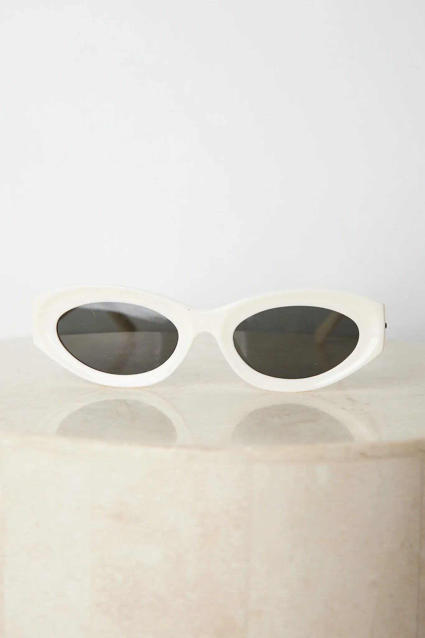 Close-up of a pair of white oval-shaped sunglasses with dark lenses, resting on a smooth beige surface against a plain white background.
