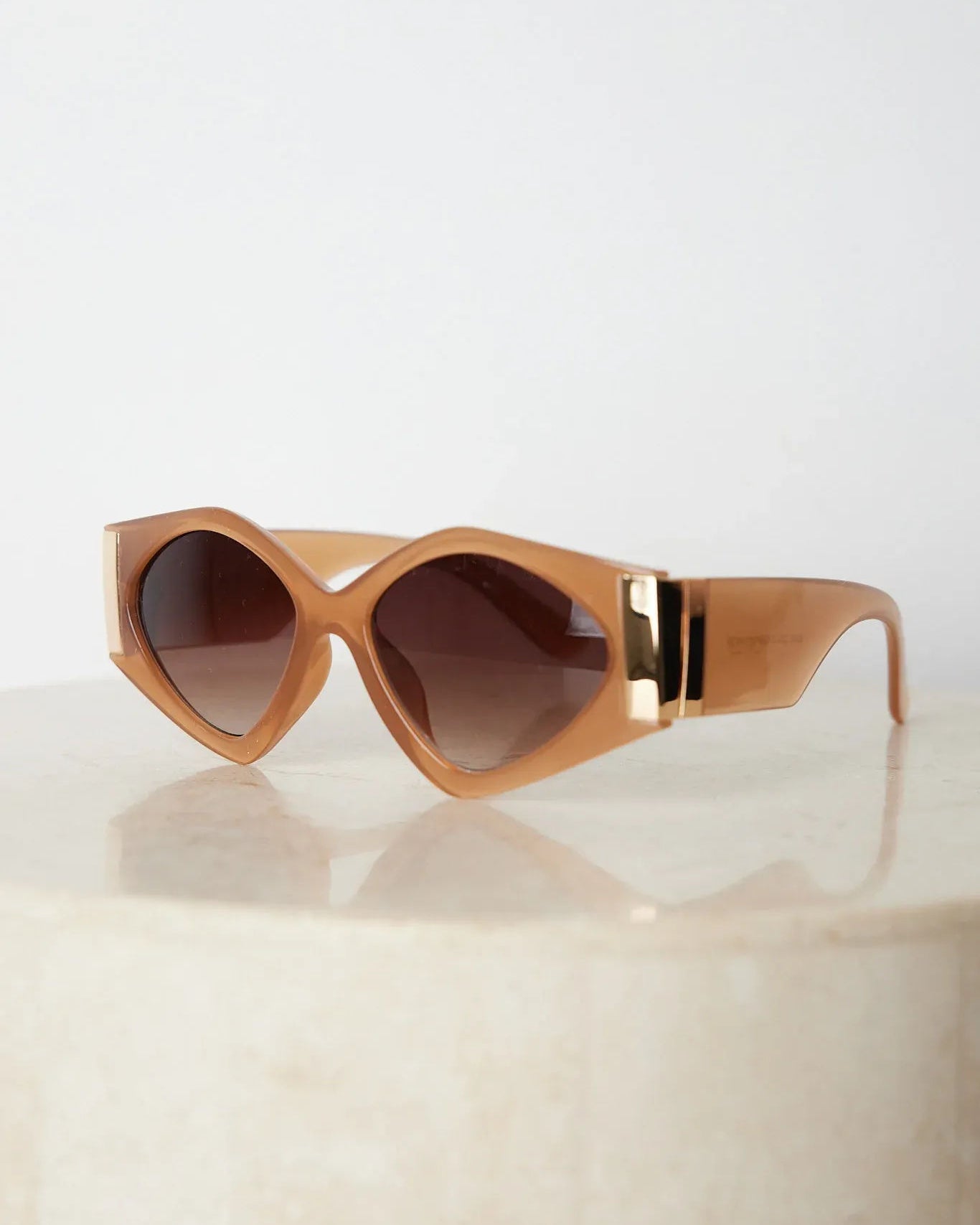 Brown cat-eye sunglasses with thick, glossy frames and dark brown tinted lenses, featuring gold accents on the corners, displayed on a light beige surface against a plain white background.
