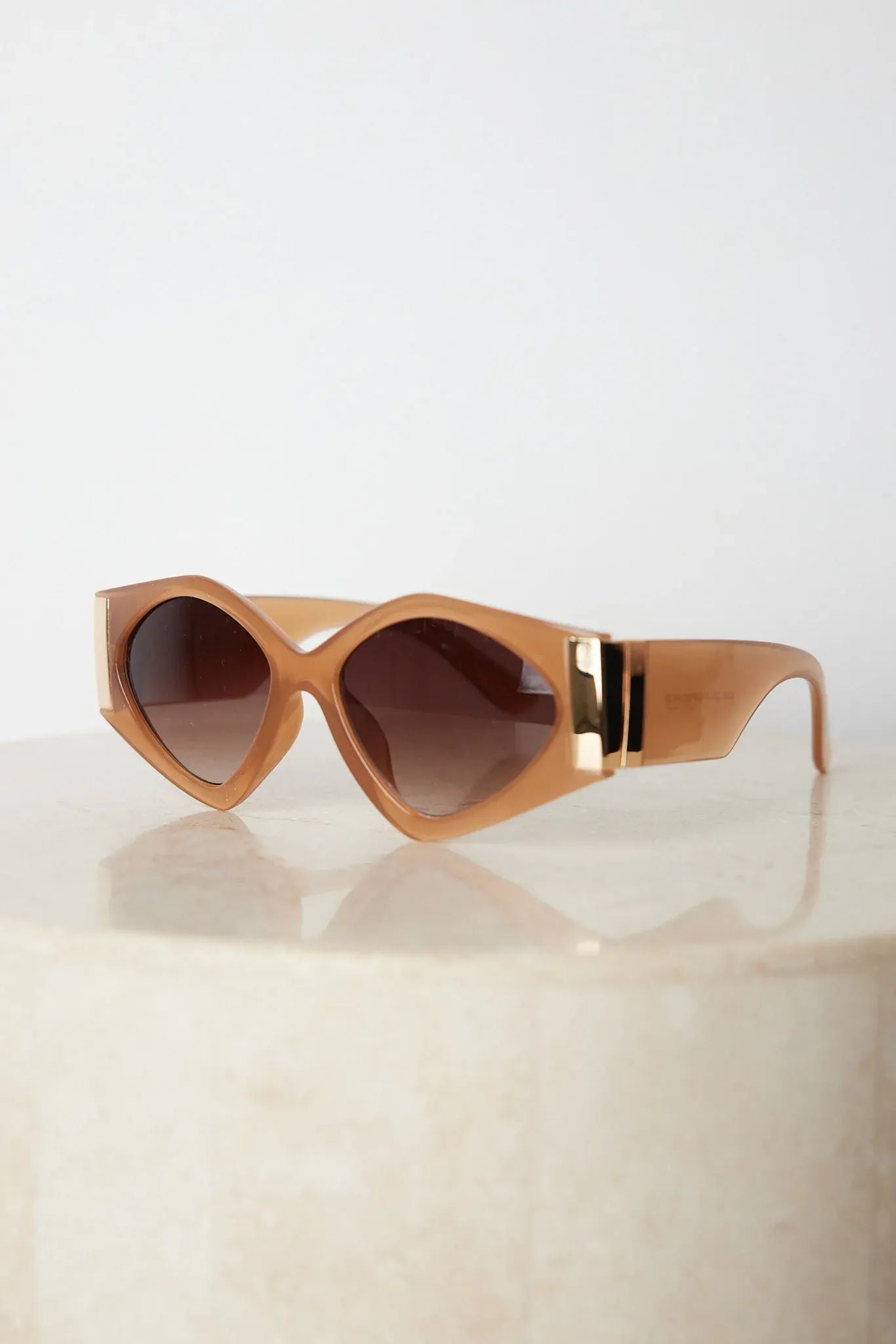 Brown cat-eye sunglasses with thick, glossy frames and dark brown tinted lenses, featuring gold accents on the corners, displayed on a light beige surface against a plain white background.