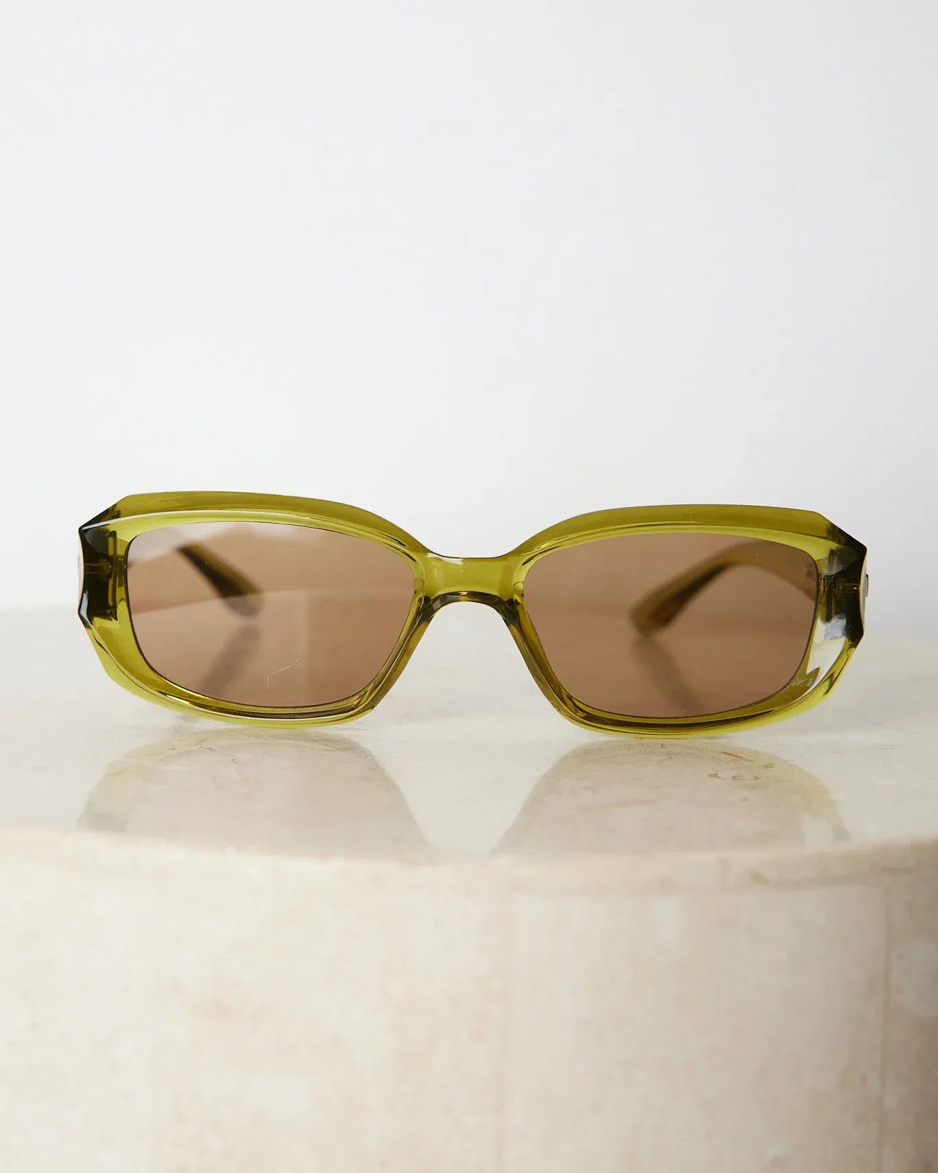 Rectangular sunglasses with translucent olive green frames and brown tinted lenses, displayed on a light beige stone surface against a plain white background.