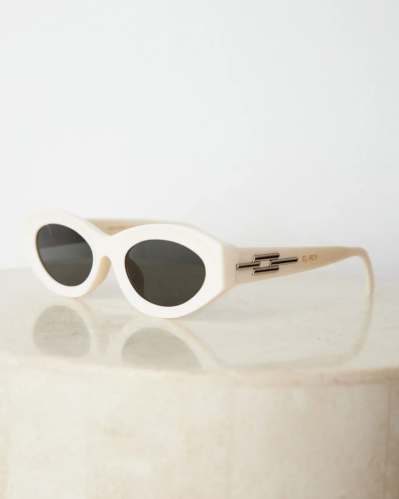 Close-up of a pair of white oval-shaped sunglasses with dark lenses, resting on a smooth beige surface against a plain white background.