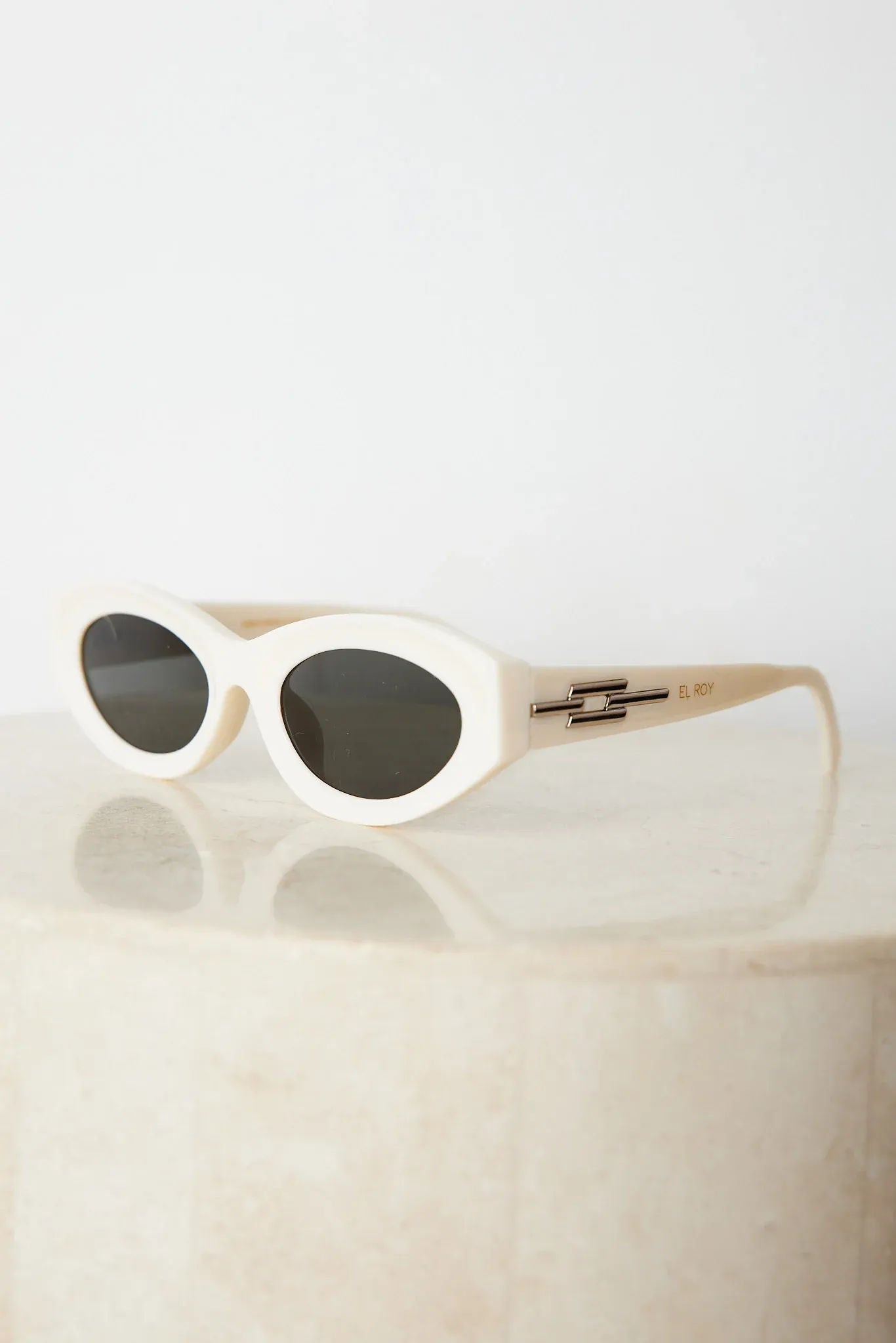 Close-up of a pair of white oval-shaped sunglasses with dark lenses, resting on a smooth beige surface against a plain white background.