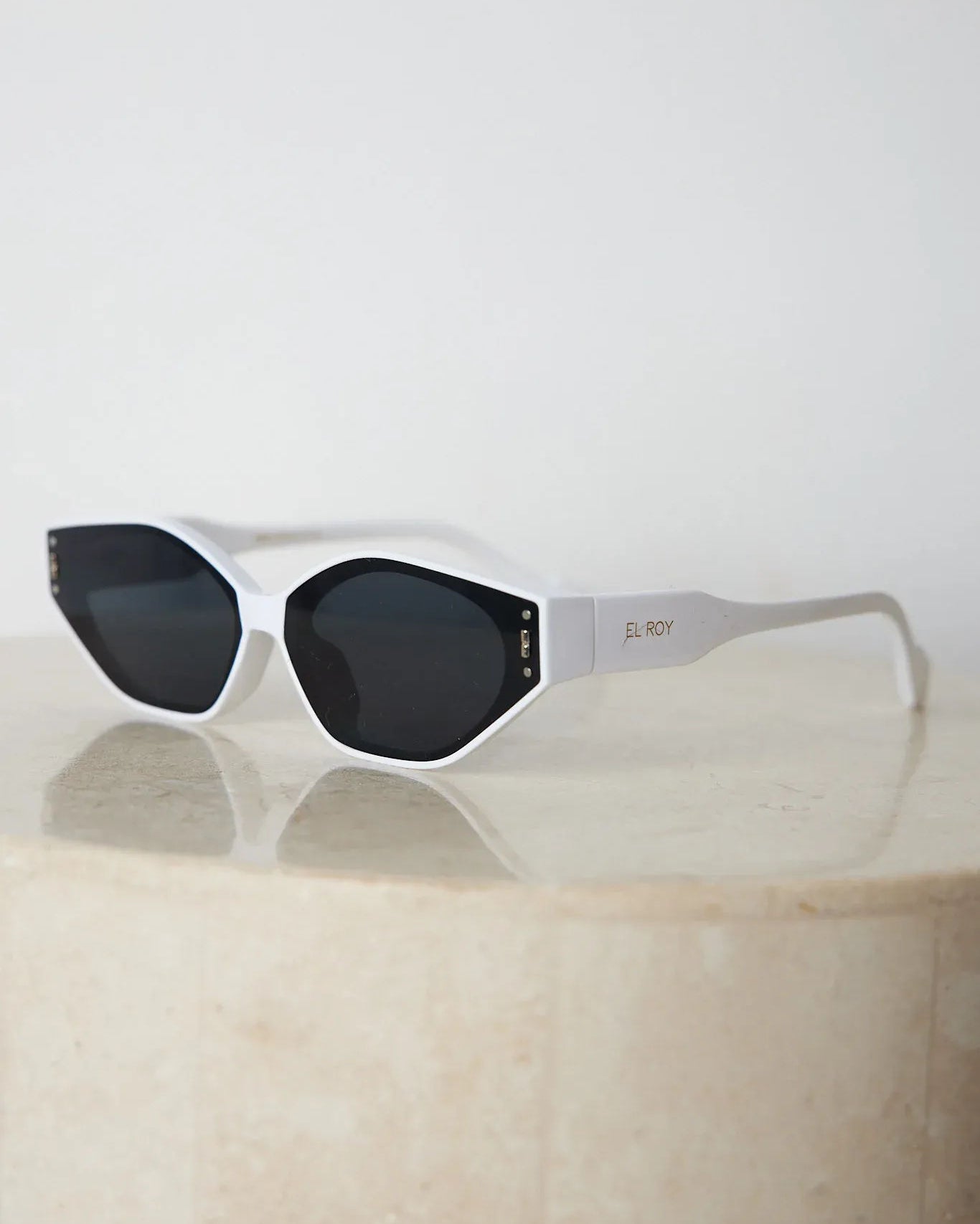 White geometric-shaped sunglasses with dark lenses resting on a light marble surface against a plain white background.