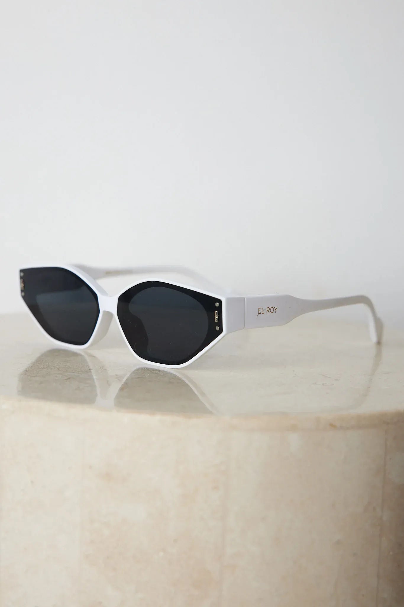 White geometric-shaped sunglasses with dark lenses resting on a light marble surface against a plain white background.