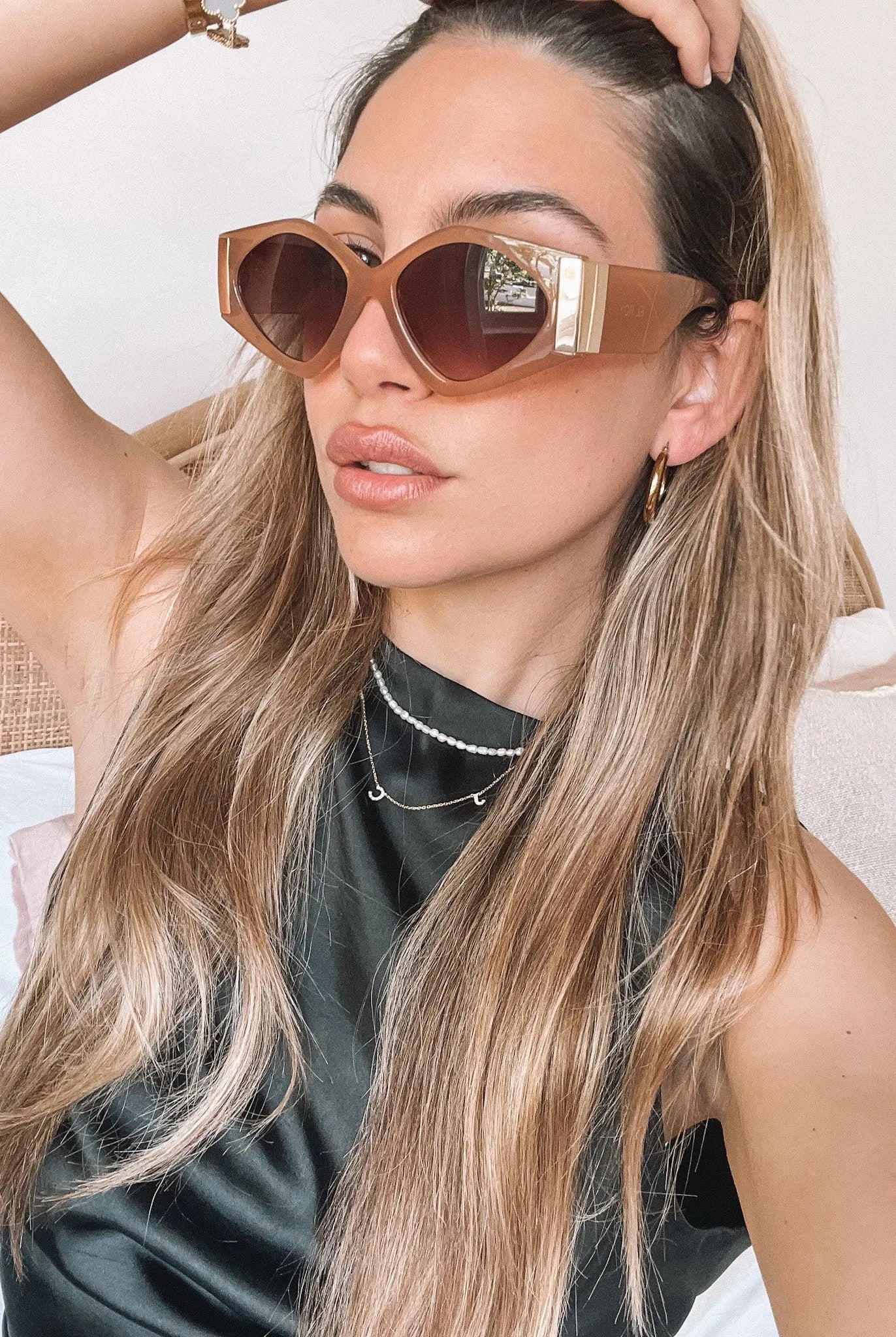 Brown cat-eye sunglasses with thick, glossy frames and dark brown tinted lenses, featuring gold accents on the corners, displayed on a light beige surface against a plain white background.