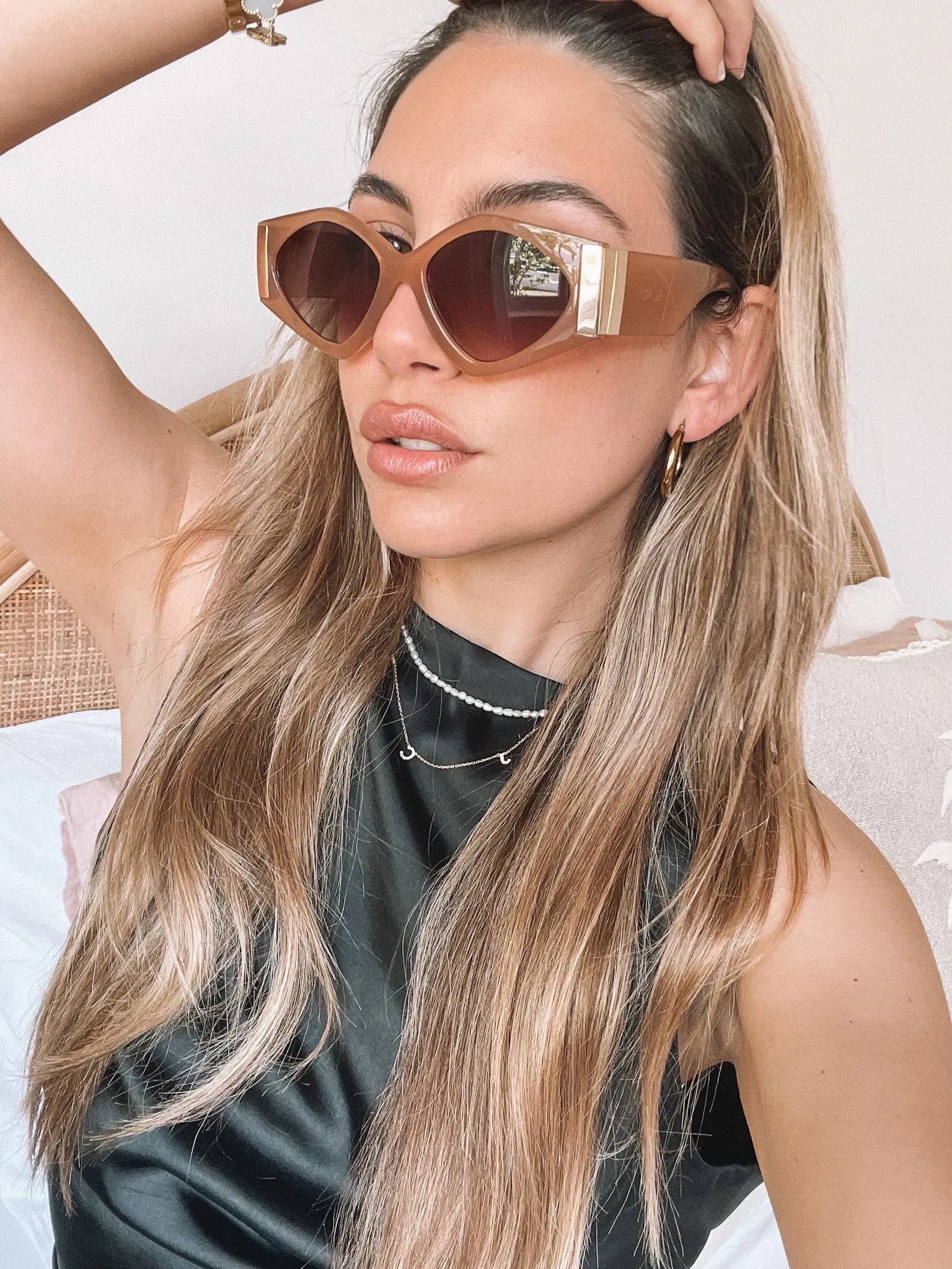 Brown cat-eye sunglasses with thick, glossy frames and dark brown tinted lenses, featuring gold accents on the corners, displayed on a light beige surface against a plain white background.