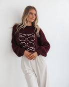 A woman with long, wavy blonde hair leans casually against a white wall, wearing a burgundy sweater with a white abstract design on the front and high-waisted cream trousers. She has her hands in her pockets and gazes confidently at the camera, set against a clean, minimalist background.