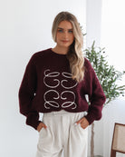 A woman with long, wavy blonde hair leans casually against a white wall, wearing a burgundy sweater with a white abstract design on the front and high-waisted cream trousers. She has her hands in her pockets and gazes confidently at the camera, set against a clean, minimalist background.