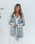 A smiling woman with long, wavy brown hair stands against a plain white background, wearing a cozy grey cardigan with white trim over a white top and loose-fitting cream pants. She has her hands gently clasped in front of her, giving a relaxed and cheerful vibe. A small sprig of greenery is visible to the left.