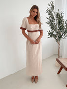 A woman with long, wavy blonde hair poses indoors in a minimalist room with white walls and a potted plant. She is wearing a light beige short-sleeve dress with brown trim details at the sleeves and waist, featuring a square neckline and a slim fit. She completes the look with pointed nude heels and a soft, confident expression.