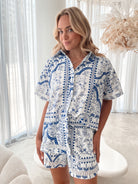 A woman with blonde, wavy hair stands in a bright, modern room with floor-to-ceiling sheer white curtains and neutral-toned decor. She is wearing a coordinated short-sleeve button-up shirt and matching shorts set featuring a blue and white floral and geometric print. She has a relaxed expression with one hand in her pocket, exuding a stylish and effortless vibe.