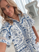 A woman with blonde, wavy hair stands in a bright, modern room with floor-to-ceiling sheer white curtains and neutral-toned decor. She is wearing a coordinated short-sleeve button-up shirt and matching shorts set featuring a blue and white floral and geometric print. She has a relaxed expression with one hand in her pocket, exuding a stylish and effortless vibe.