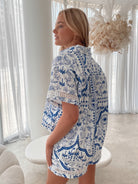 A woman with blonde, wavy hair stands in a bright, modern room with floor-to-ceiling sheer white curtains and neutral-toned decor. She is wearing a coordinated short-sleeve button-up shirt and matching shorts set featuring a blue and white floral and geometric print. She has a relaxed expression with one hand in her pocket, exuding a stylish and effortless vibe.