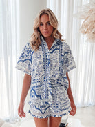 A woman with blonde, wavy hair stands in a bright, modern room with floor-to-ceiling sheer white curtains and neutral-toned decor. She is wearing a coordinated short-sleeve button-up shirt and matching shorts set featuring a blue and white floral and geometric print. She has a relaxed expression with one hand in her pocket, exuding a stylish and effortless vibe.