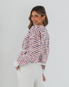 A woman with long, wavy brown hair stands against a plain white background, wearing a textured cardigan with a red, pink, and navy checkered pattern. The cardigan features large gold buttons and front pockets, layered over a white knit top. She pairs it with light cream-colored pants and poses with her hands in her pockets, gazing slightly to the side with a composed expression.