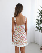 A woman wearing a sleeveless white mini dress with a scalloped hem and floral lattice pattern in pink, yellow, and green tones. The dress features wide crocheted straps and a sweetheart neckline. She stands in a minimalistic room with white walls and a potted plant in the background.