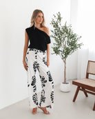 A woman sitting on a wooden chair wearing a black off-the-shoulder top paired with high-waisted white wide-leg pants featuring bold black tropical leaf prints. She completes the look with nude heels and is posed in a minimalistic room with white curtains and a potted plant in the background.