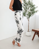 A woman sitting on a wooden chair wearing a black off-the-shoulder top paired with high-waisted white wide-leg pants featuring bold black tropical leaf prints. She completes the look with nude heels and is posed in a minimalistic room with white curtains and a potted plant in the background.