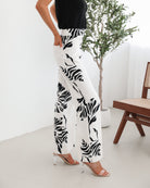 A woman sitting on a wooden chair wearing a black off-the-shoulder top paired with high-waisted white wide-leg pants featuring bold black tropical leaf prints. She completes the look with nude heels and is posed in a minimalistic room with white curtains and a potted plant in the background.