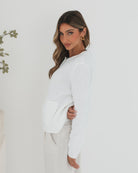 A woman wearing a white textured knit cardigan with front buttons and two small front pockets, paired with high-waisted light beige trousers. She stands against a plain white background, with one hand in her pocket and the other touching her hair.