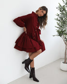 Smiling woman with long wavy hair wearing a loose-fitting, deep red button-up dress with ruffled tiers and three-quarter sleeves, paired with black ankle boots. She is standing indoors on a light-colored floor, holding the hem of her dress with one hand. A potted green plant and a wooden chair are visible in the background.